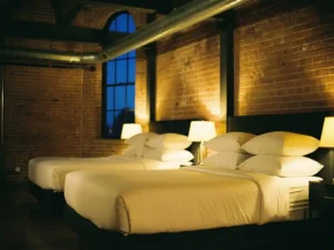 Hotelkamerbedden met wit linnengoed in industriële loft met bakstenen muren, stalen balken en sfeervol lamplicht bij avondschemering.