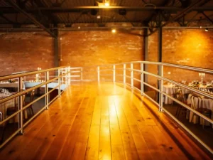 Houten rolstoelhelling met gepolijste metalen leuningen in industriële evenementenhal met bakstenen muren en gedekte tafels op de achtergrond.