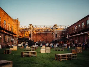 Industrieel festivalterrein met hoofdpodium tussen bakstenen magazijnmuren, groen veld en sfeervol slingerverlichting in gouden avondlicht.