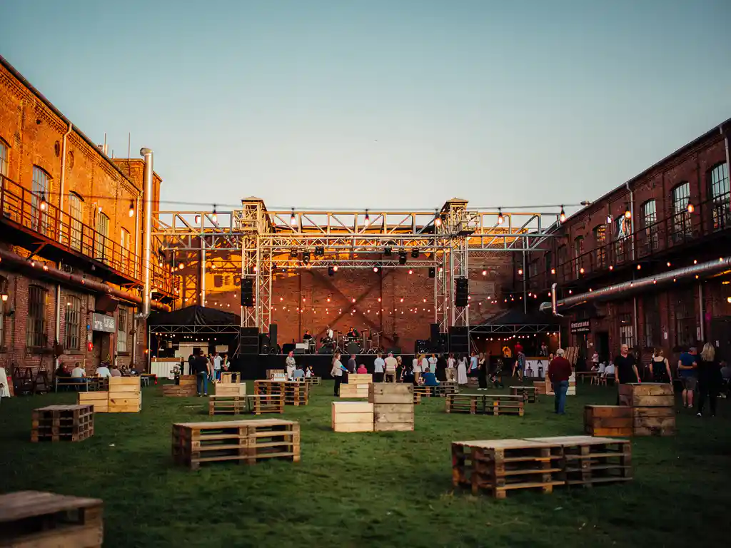 Industrieel festivalterrein met hoofdpodium tussen bakstenen magazijnmuren, groen veld en sfeervol slingerverlichting in gouden avondlicht.