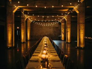 Industriële feestzaal met amberkleurige uplighting op betonnen pilaren, Edison-peerlampjes en gouden kaarslicht op lange banquettafels.