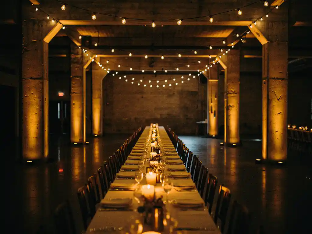Industriële feestzaal met amberkleurige uplighting op betonnen pilaren, Edison-peerlampjes en gouden kaarslicht op lange banquettafels.