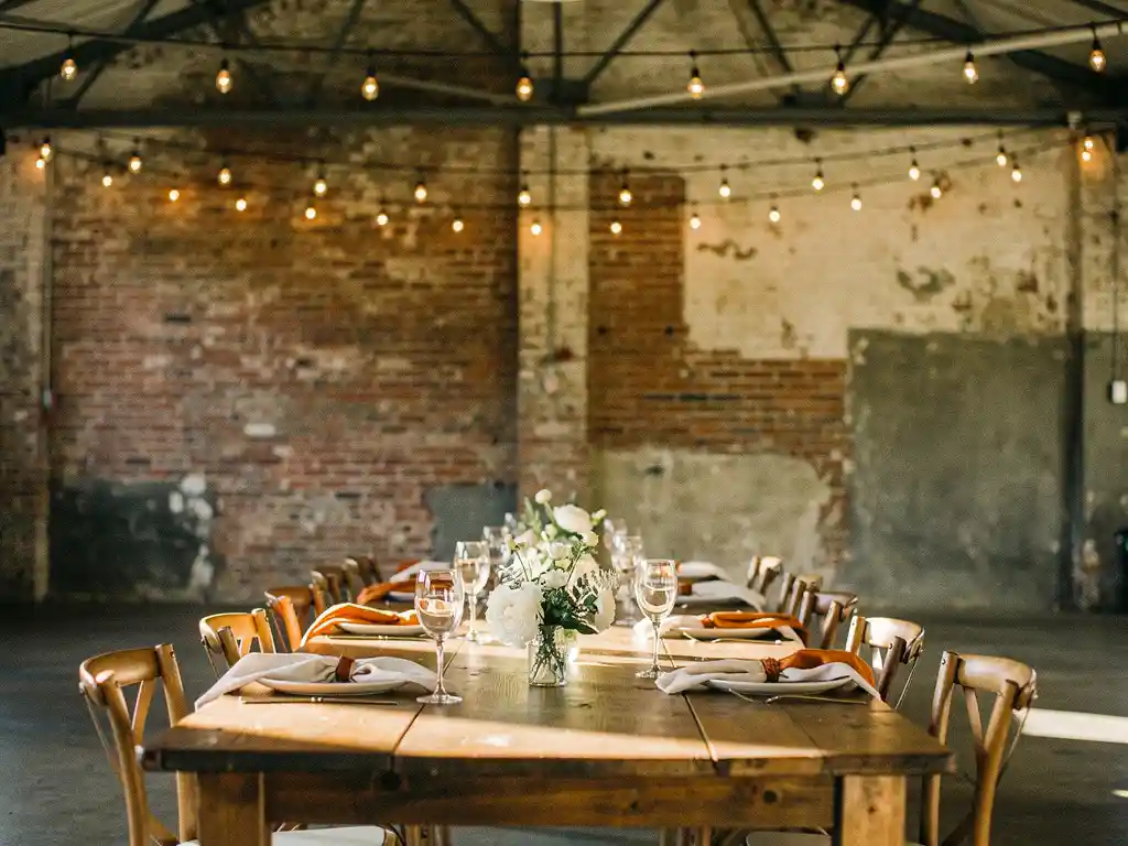 Stijlvol gedekte evenementtafel met elegant glaswerk en bloemstukken in industriële bakstenen zaal met sfeervolle Edison-peertjes.