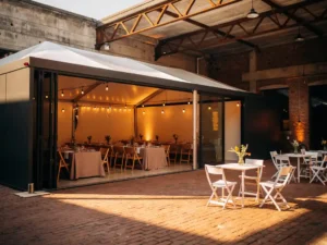 Moderne evenementenhal met Edison-verlichting en elegante tafels naast een rustieke pop-uptent op bakstenen binnenplaats in gouden middaglicht.