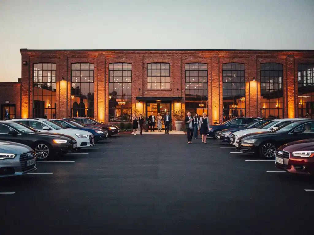 Gasten lopen bij schemering naar een industrieel bakstenen evenementenlocatie met verlichte ramen en volle parkeerplaats.